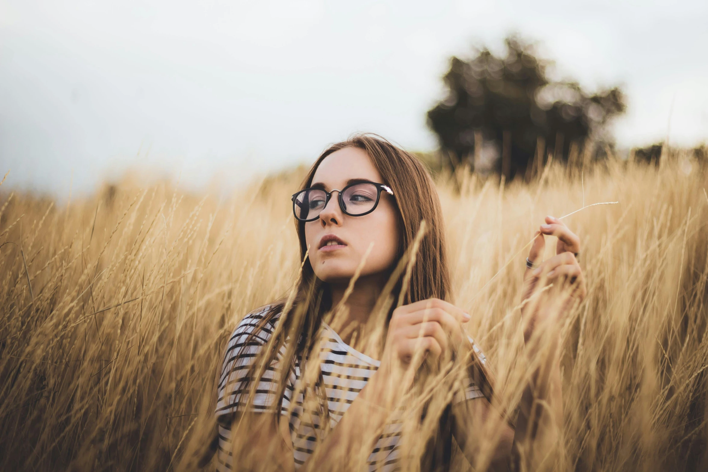 a woman standing in a field of tall grass, trending on pexels, square rimmed glasses, beautiful lonely girl, avatar image, straw