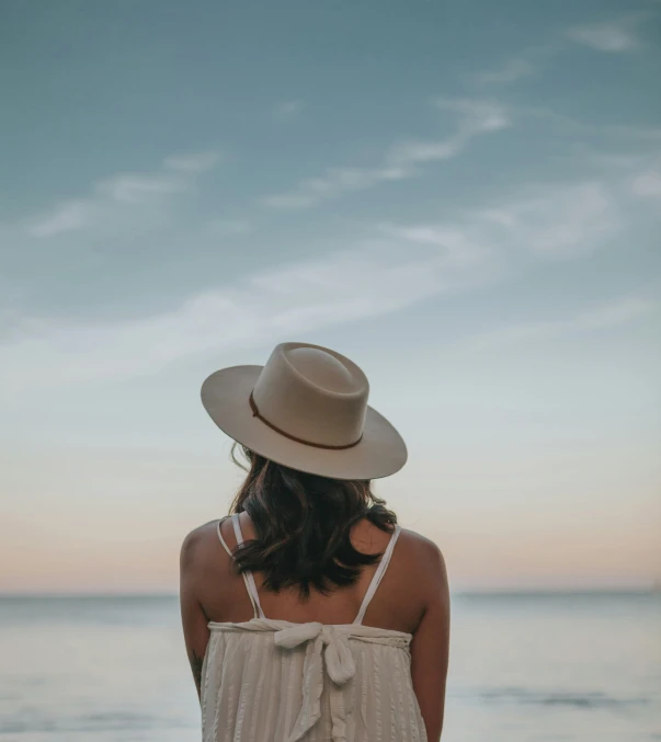 a woman standing on a beach looking out at the ocean, pexels contest winner, romanticism, beige fedora, summer evening, simple muted colors, pictured from the shoulders up