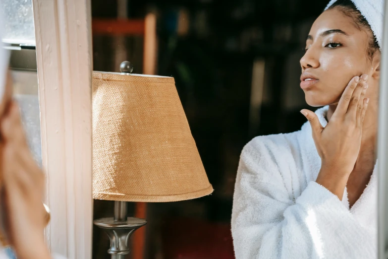 a woman in a bathrobe looking at her reflection in a mirror, trending on pexels, emerging from her lamp, with a pointed chin, warm sunshine, skincare