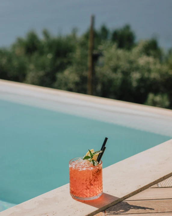 a drink sitting on top of a wooden table next to a pool, pexels contest winner, renaissance, lgbtq, greek setting, with a straw, cosmopolitan