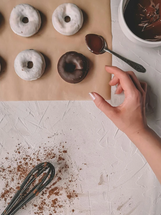 a person holding a whisk next to doughnuts on a table, by Robbie Trevino, trending on pexels, chocolate art, partially cupping her hands, detailed product image, shot from a drone