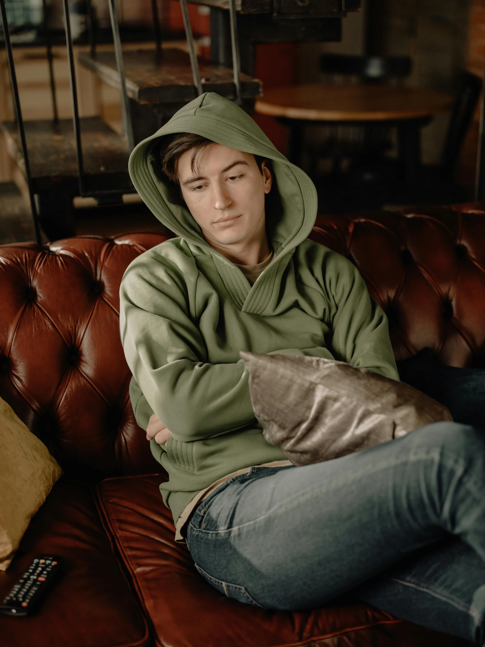 a man sitting on a couch wearing a green hoodie, a colorized photo, by Elsa Bleda, trending on unsplash, levi ackerman, around 1 9 years old, slightly tanned, frown fashion model