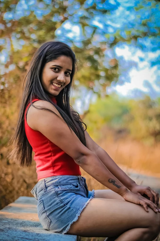 a woman sitting on top of a wooden bench, by Max Dauthendey, pexels contest winner, realism, indian girl with brown skin, long hair and red shirt, wearing a camisole and shorts, while smiling for a photograph
