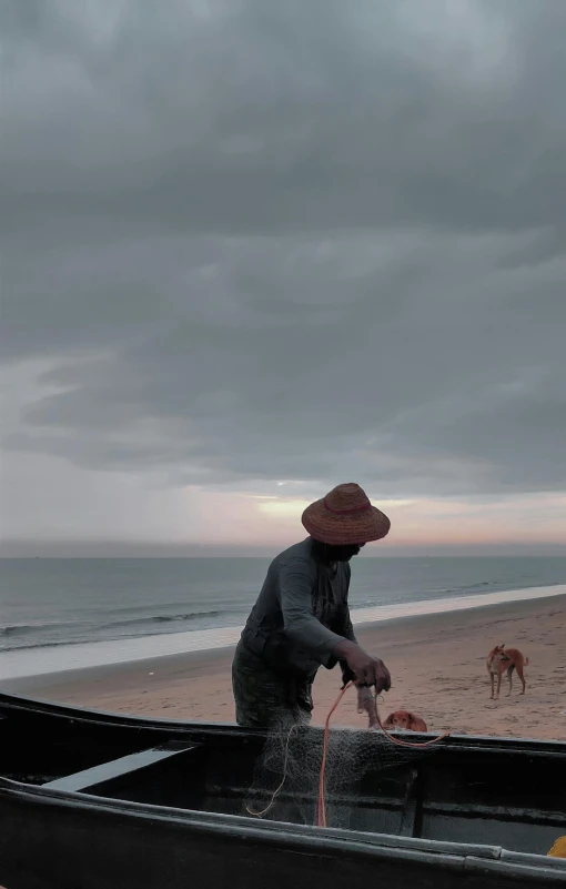 a man standing next to a boat on a beach, inspired by Steve McCurry, unsplash, fantastic realism, low quality footage, with stray dogs, overcast!!! cinematic focus, panoramic shot