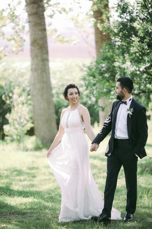 a man in a tuxedo and a woman in a wedding dress holding hands, walking through the trees, greek ameera al taweel, in a open green field, smiling couple