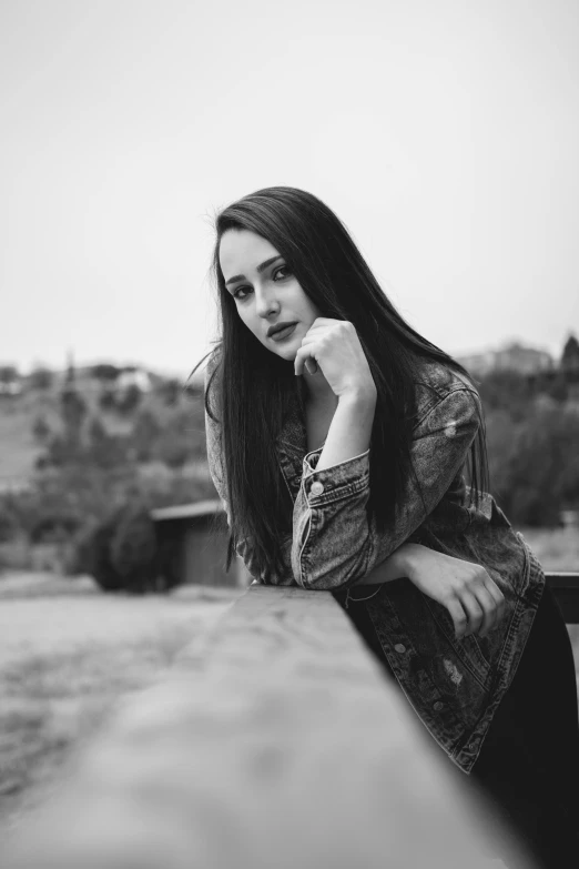 a black and white photo of a woman leaning on a wall, a black and white photo, pexels contest winner, young woman with long dark hair, sitting on bench, hills in the background, gray skin. grunge