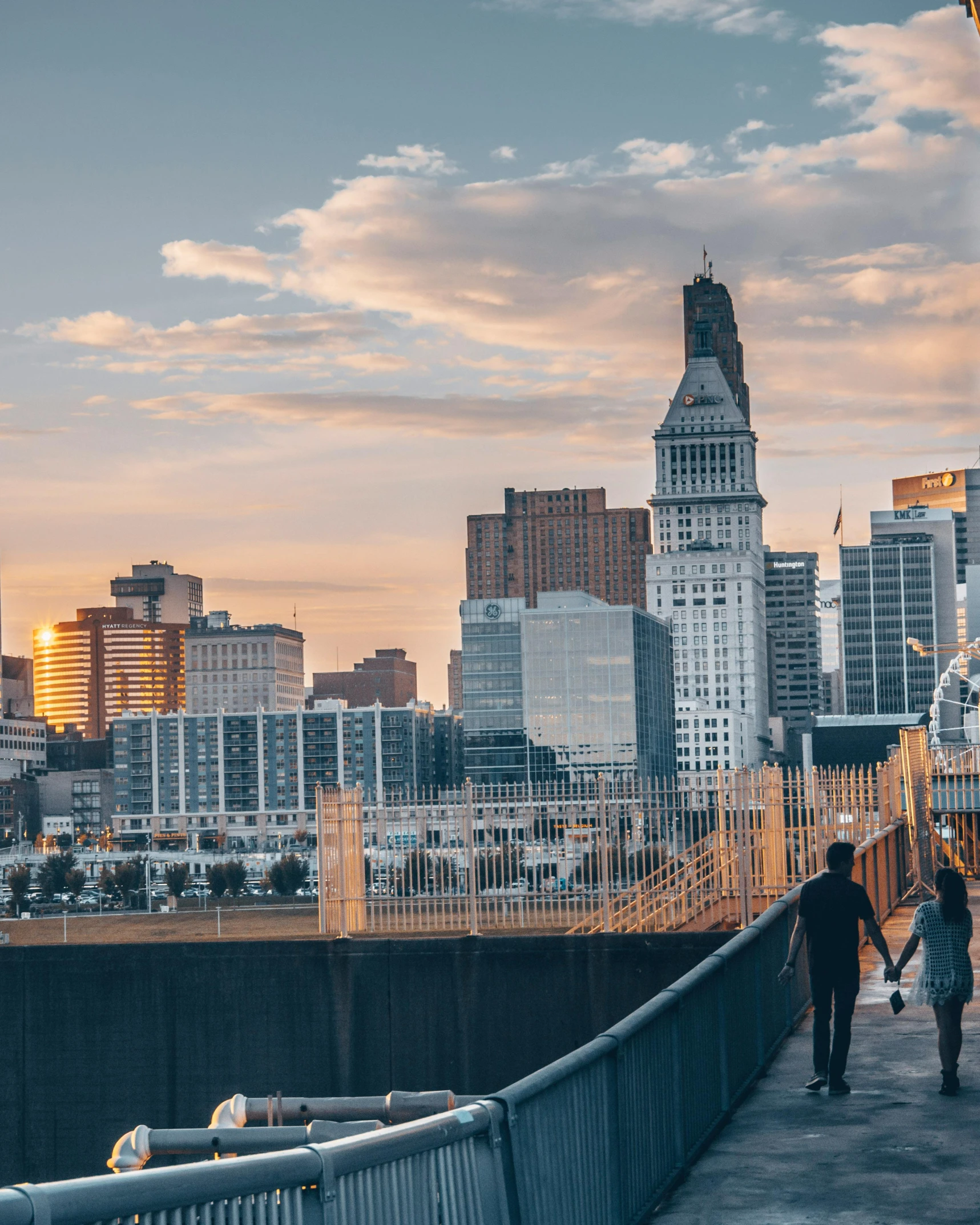 a couple of people walking across a bridge, by Jacob Burck, unsplash contest winner, graffiti, downtown in the distance, cleveland, vista of a city at sunset, lgbtq