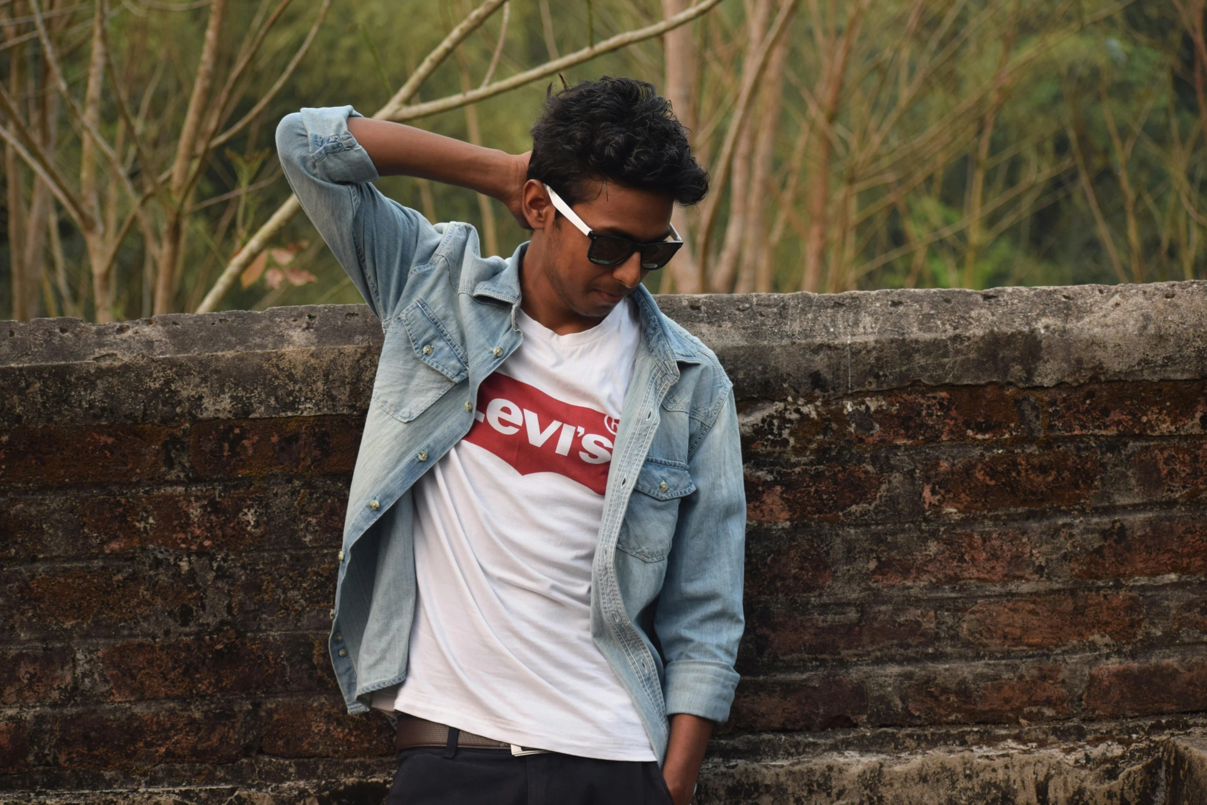 a man leaning against a brick wall smoking a cigarette, a picture, inspired by Narayan Shridhar Bendre, unsplash, graphic tees, wear ray - ban glass, vintage levi ’ s ad, eyelevel!!! view!!! photography