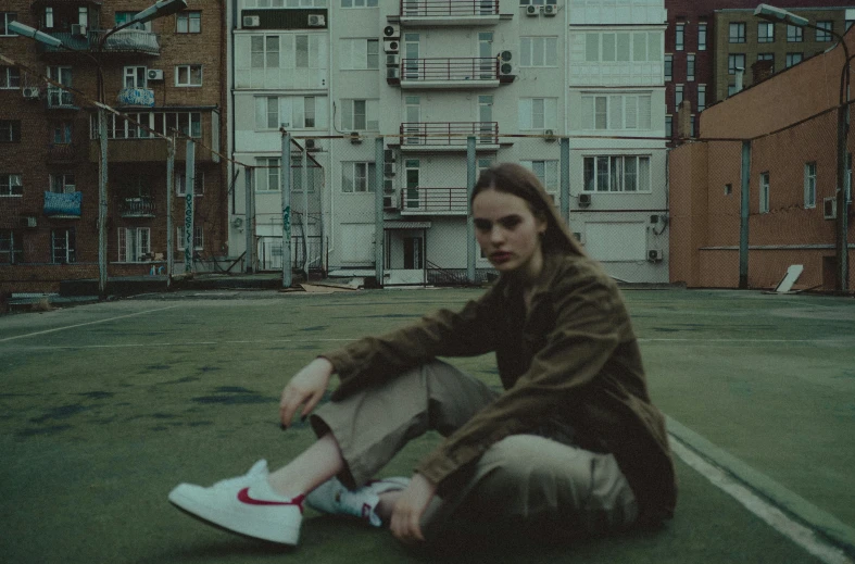 a woman sitting on the ground in front of a building, an album cover, by Attila Meszlenyi, pexels contest winner, realism, sneaker, portrait androgynous girl, grainy movie still, greta thunberg