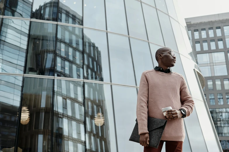 a man standing in front of a tall building, inspired by Harrington Mann, pexels contest winner, adut akech, brown and pink color scheme, canary wharf, wearing an oversized sweater