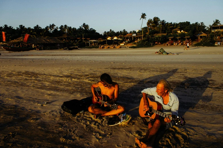 a couple of men sitting on top of a sandy beach, playing guitars, zac retz, india, lush surroundings