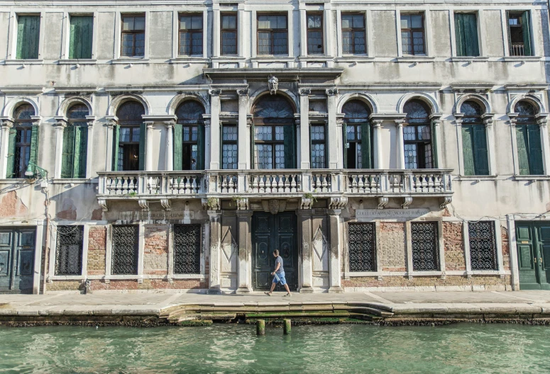 a person walking in front of a building next to a body of water, by Canaletto, pexels contest winner, many large green windows, vogue italy, payne's grey and venetian red, mansion