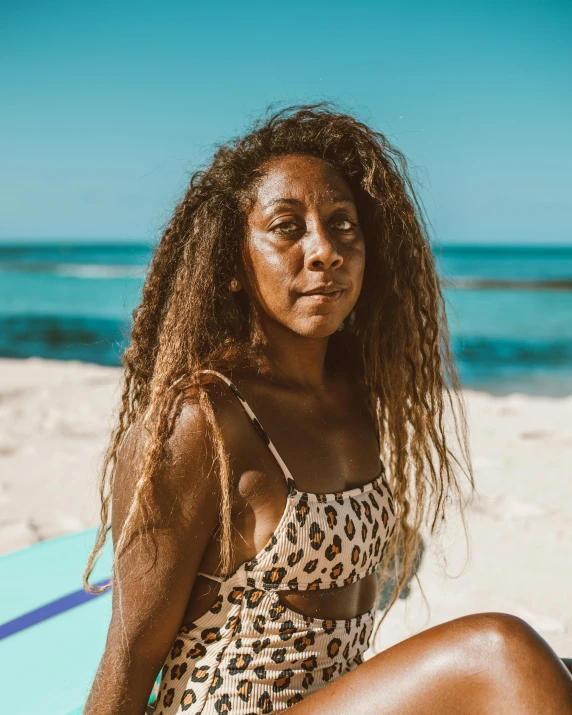 a woman sitting on top of a surfboard on a beach, by Olivia Peguero, trending on unsplash, afrofuturism, with textured hair and skin, looking straight into camera, slightly tanned, with freckles