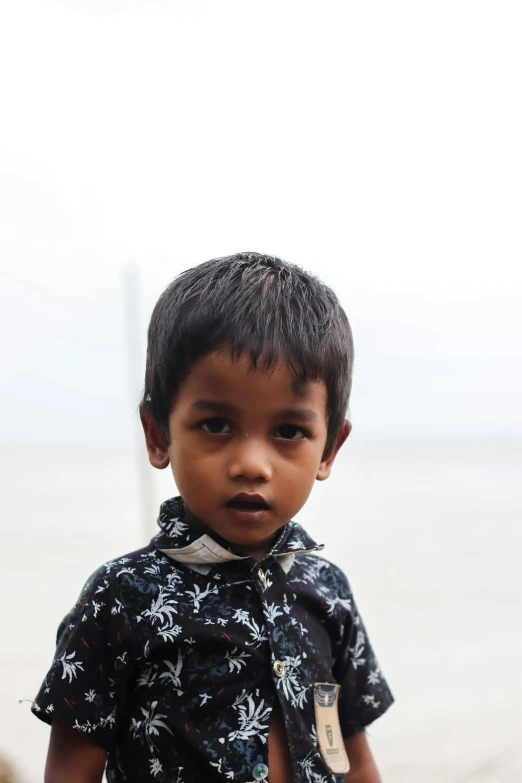 a young boy standing on a beach next to the ocean, by Basuki Abdullah, boyish face, on a gray background, assamese, small ears