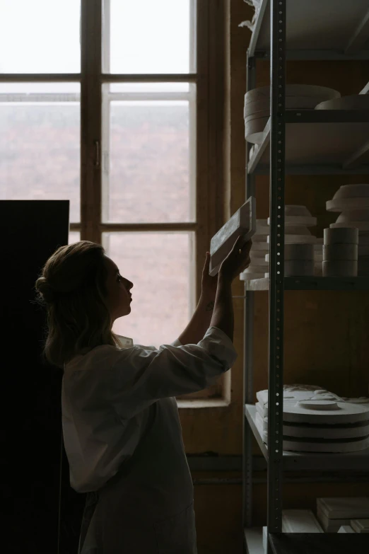 a woman that is standing in front of a window, shelves, plating, dusty light, ceramic