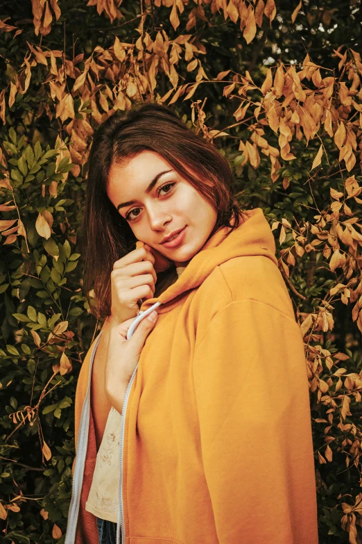 a beautiful young woman standing in front of a bush, a picture, inspired by Elsa Bleda, trending on pexels, renaissance, long orange sweatshirt, charli xcx, yellow robe, portait photo profile picture