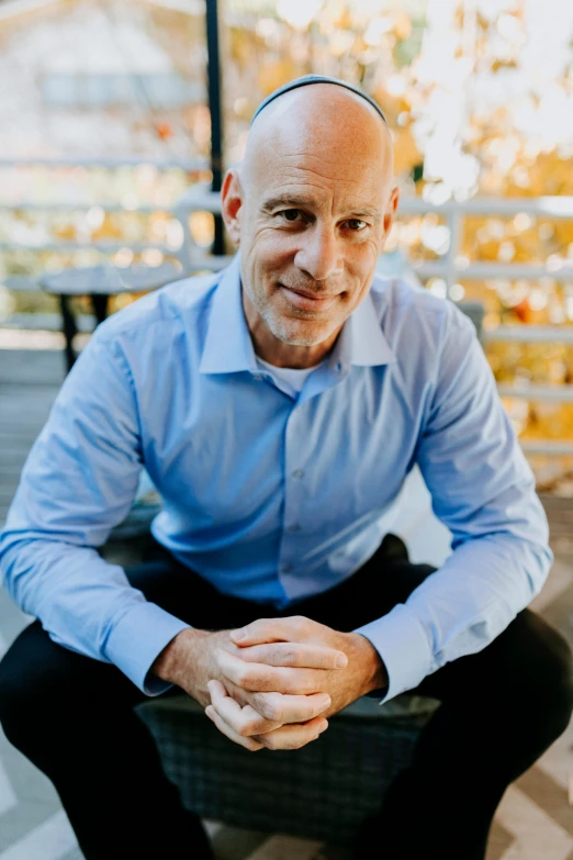 a man in a blue shirt and black pants, bald on top, vp of marketing, sitting with wrists together, adi meyers