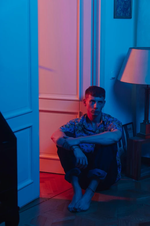 a man sitting on the floor in a room, an album cover, low saturated red and blue light, non binary model, annoyed, ignant