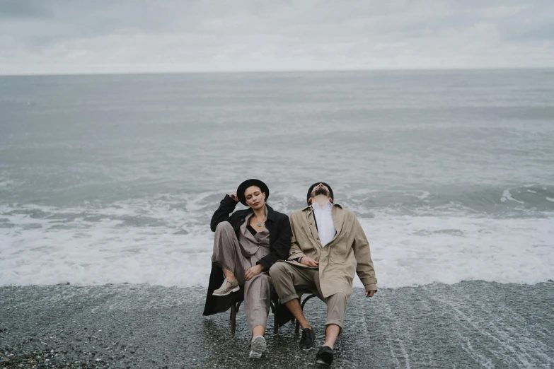 a couple of people sitting on top of a beach, by Emma Andijewska, unsplash, surrealism, wearing a trenchcoat, lesbian, background image, gray