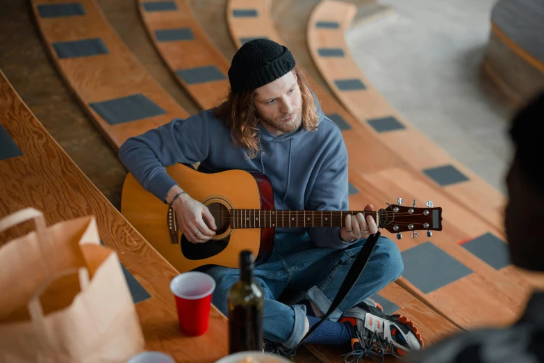 a man sitting on the ground playing a guitar, holding a drink, mini amphitheatre, student, indoor setting