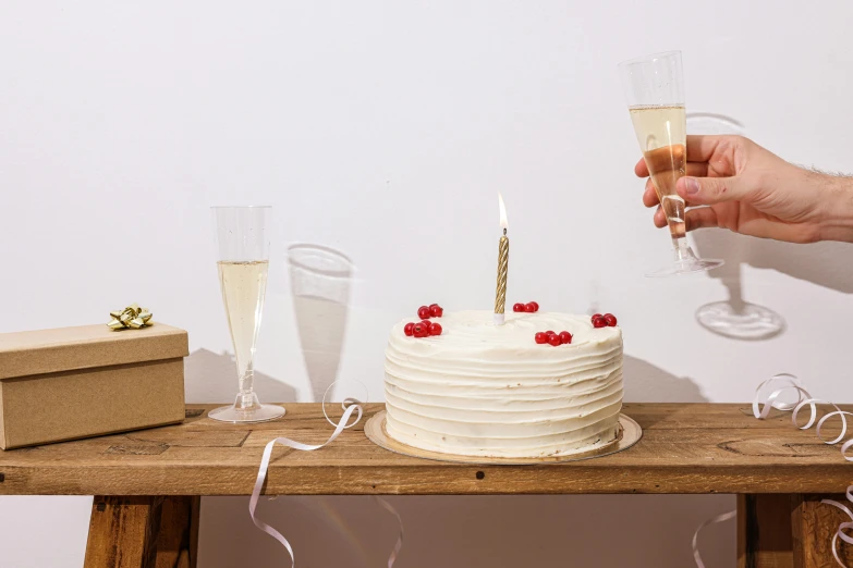 a person holding a glass of champagne in front of a cake, a still life, trending on unsplash, figuration libre, background image, birthday cake on the ground, the candle is on a wooden table, john pawson