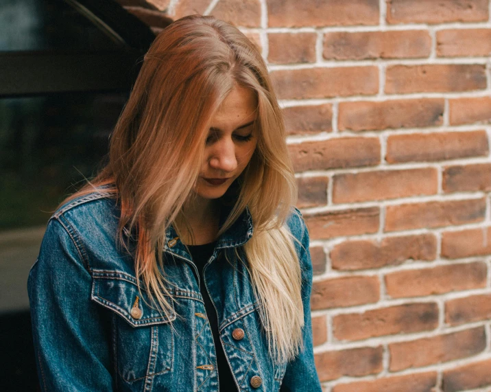 a woman standing in front of a brick building, inspired by Elsa Bleda, trending on pexels, realism, denim jacket, dark blonde hair, shy looking down, 15081959 21121991 01012000 4k