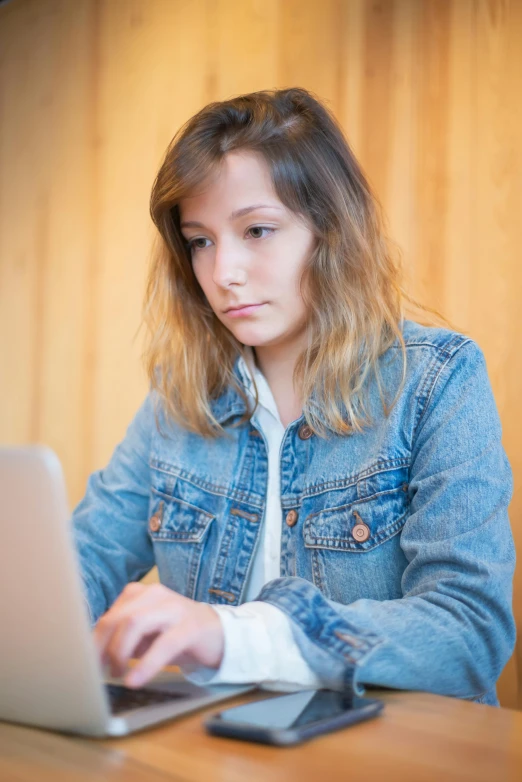 a woman sitting at a table using a laptop computer, wearing a blue jacket, serious composure, photo of the girl, teenage