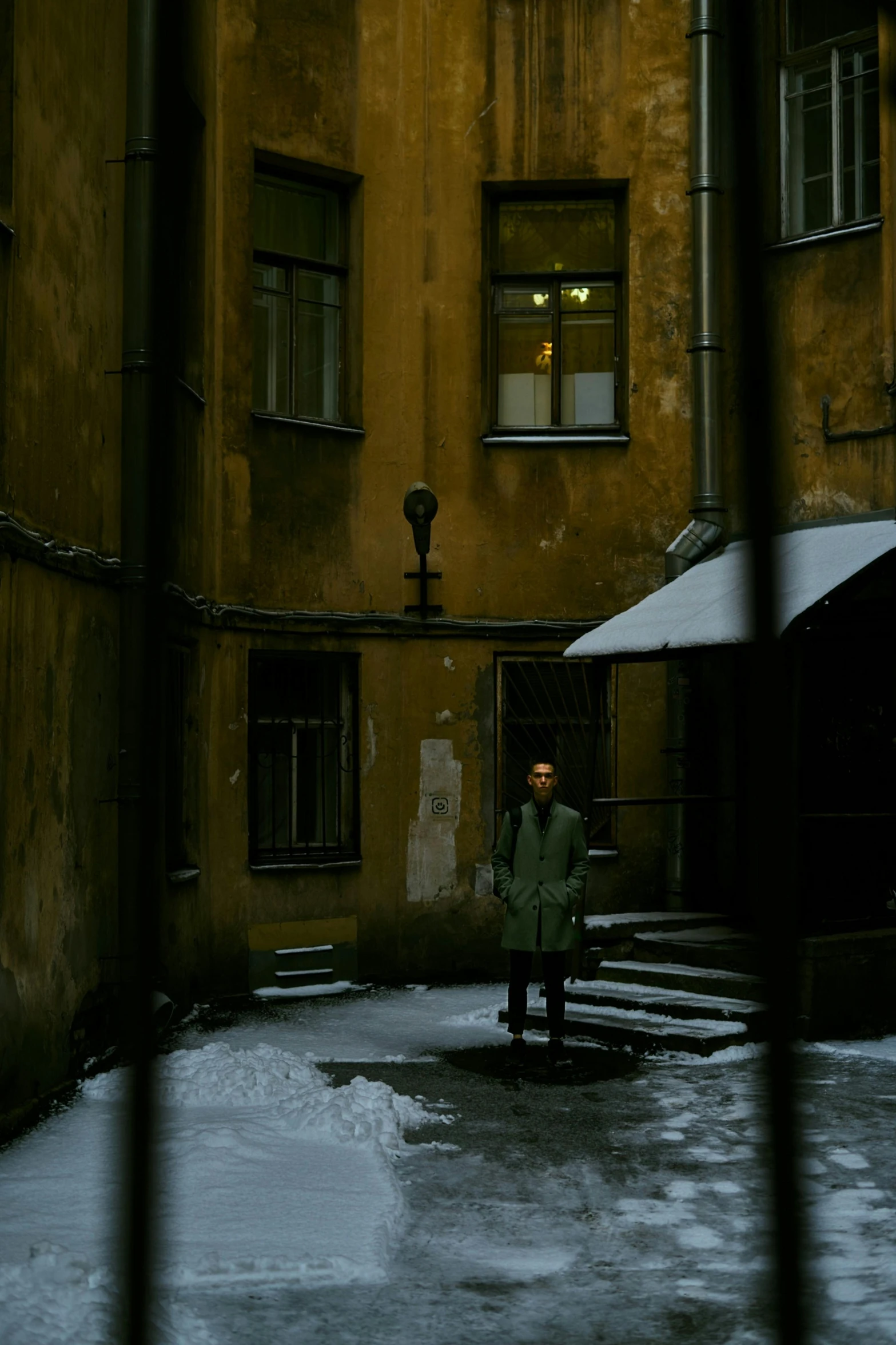 a person standing outside of a building in the snow, an album cover, inspired by Elsa Bleda, pexels contest winner, post - soviet courtyard, dark shadowy surroundings, movie still 8 k, green alleys