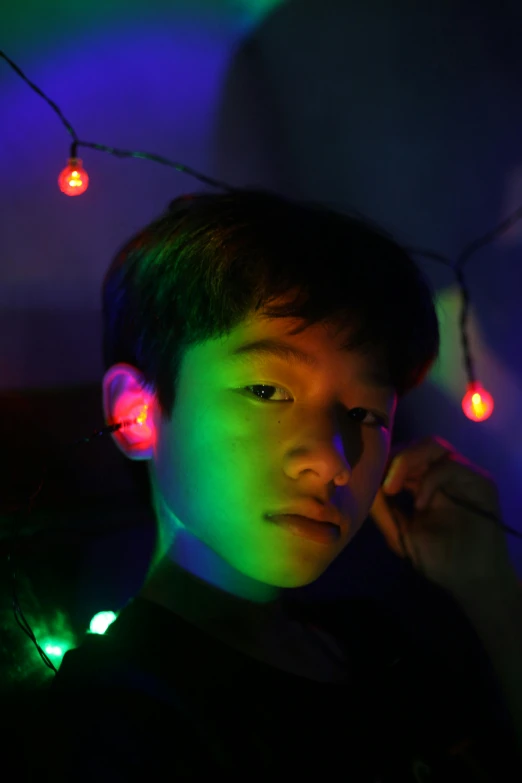 a young man standing in front of a wall covered in christmas lights, by Byron Galvez, pexels contest winner, young cute wan asian face, blue and green and red tones, aged 13, discord profile picture