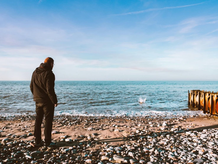 a man standing on top of a beach next to the ocean, a picture, unsplash, visual art, brown, profile pic, devastated, seaside