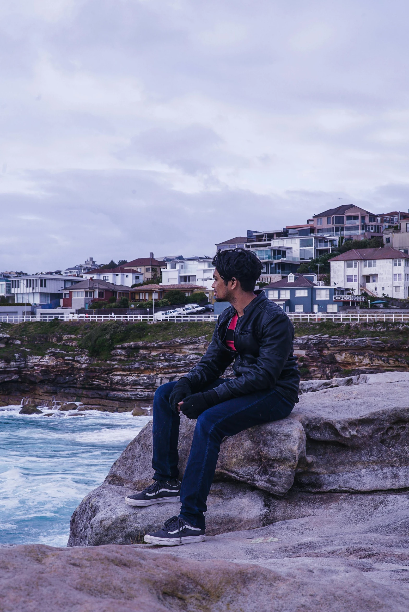 a man sitting on top of a rock next to the ocean, inspired by Sydney Carline, pexels contest winner, city backdrop, wearing jeans and a black hoodie, profile image, bondi beach in the background