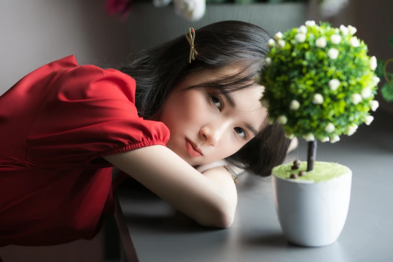 a woman leaning on a table next to a potted plant, inspired by Yukimasa Ida, pexels contest winner, girl wears a red dress, china doll face, beautiful grumpy girl, young cute wan asian face