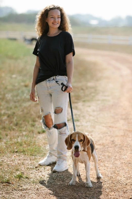 a woman walking a dog down a dirt road, ripped jeans, standing in an arena, jeans and t shirt, cute beagle