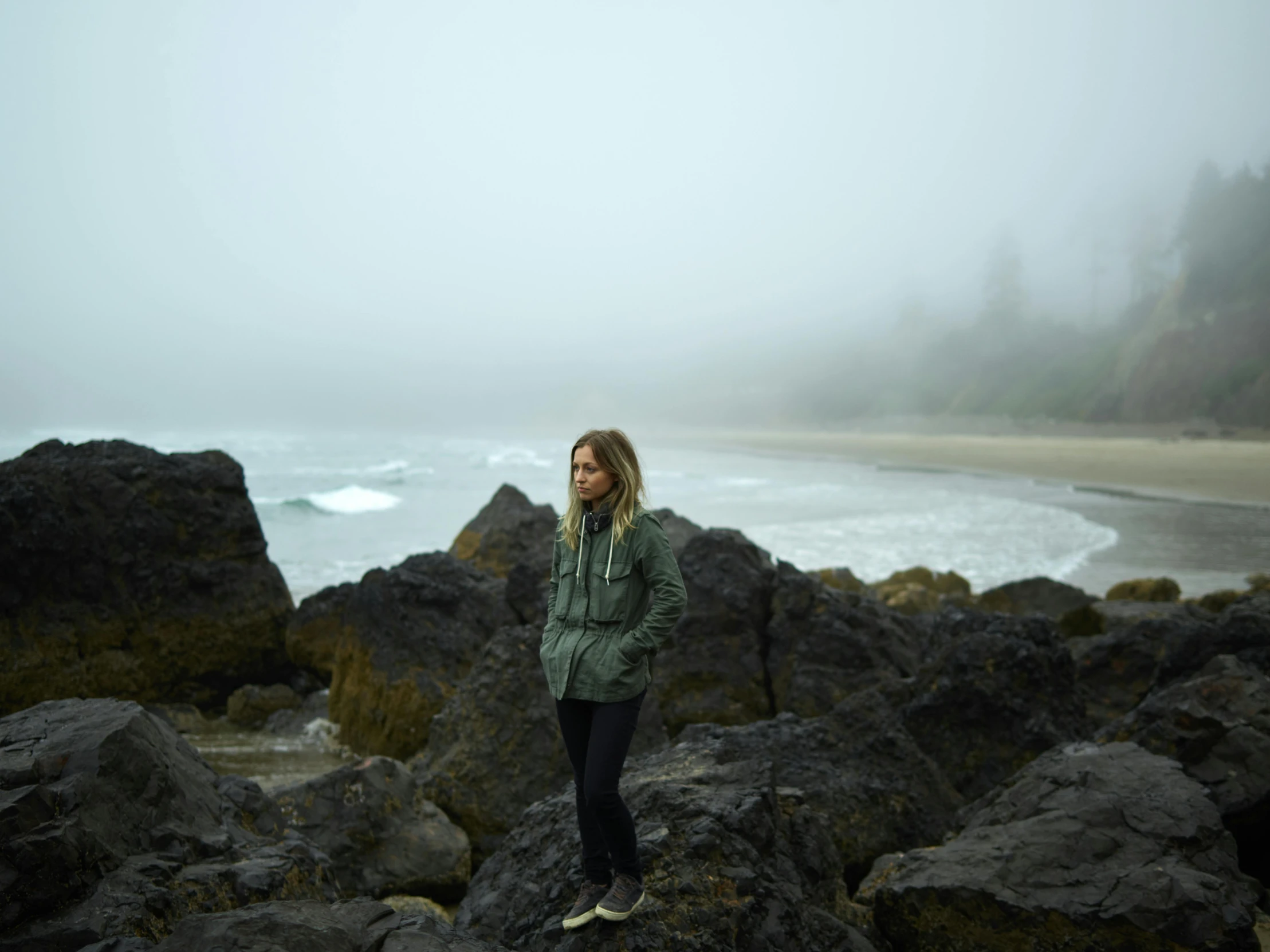 a woman standing on a rocky beach next to the ocean, wearing jacket, sydney sweeney, in the thick fog, guide