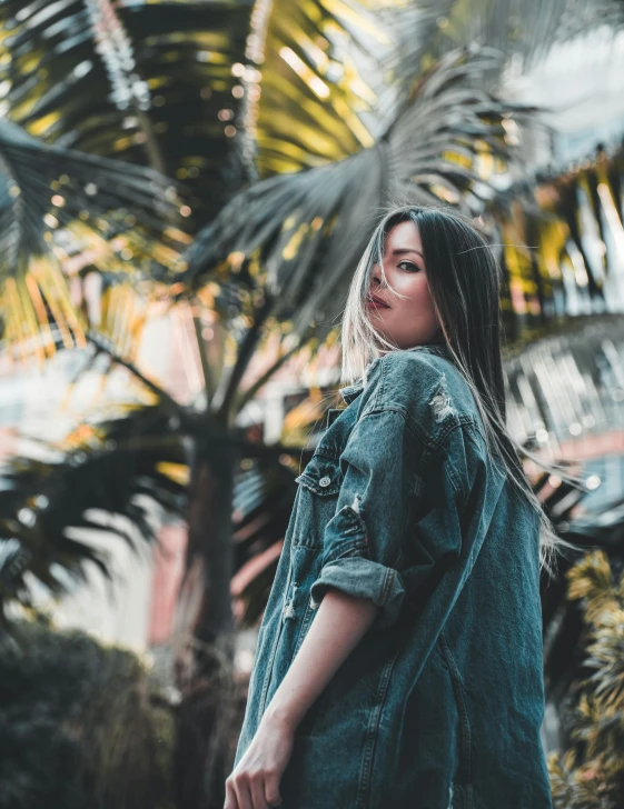 a woman standing in front of a palm tree, inspired by Elsa Bleda, trending on unsplash, sumatraism, denim jacket, low quality photo, wearing a grey robe, attractive photo