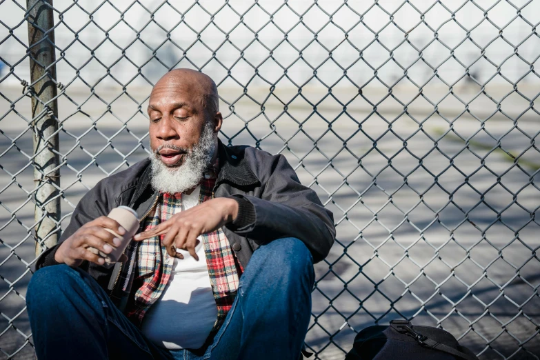 a man sitting in front of a chain link fence, by Washington Allston, pexels contest winner, bald head and white beard, jamal campbell, on the concrete ground, begging