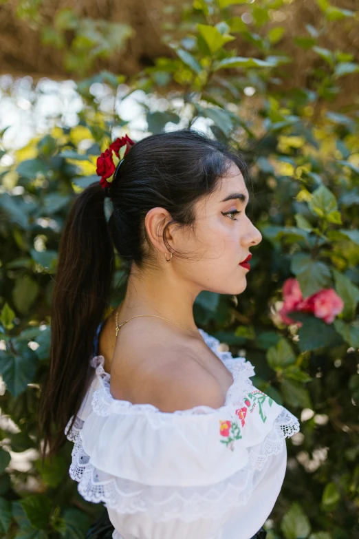 a woman in a white dress standing in front of a bush, an album cover, inspired by Frida Kahlo, pexels, black ponytail hair, profile image, red adornments, isabela moner