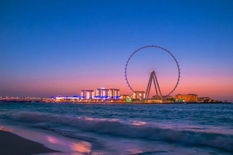 a ferris wheel sitting on top of a beach next to the ocean, ameera al taweel, vista of a city at sunset, in the center of the image