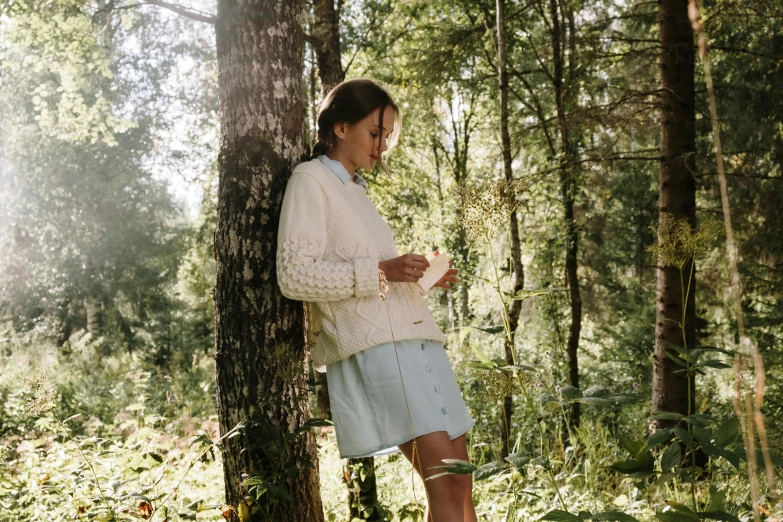 a woman standing next to a tree in a forest, unsplash, figuration libre, village girl reading a book, wearing a white sweater, letterboxing, three - quarter view