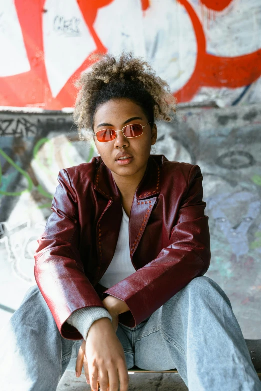 a woman sitting in front of a graffiti covered wall, an album cover, inspired by Esaias Boursse, trending on unsplash, red leather jacket, amber glasses, mixed-race woman, portrait androgynous girl