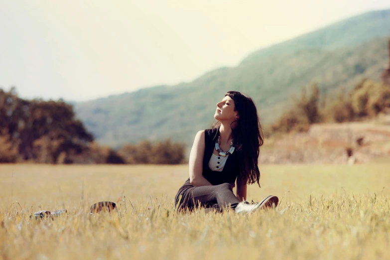 a woman sitting in a field with mountains in the background, a picture, profile image, vintage vibe, young woman looking up, very sunny