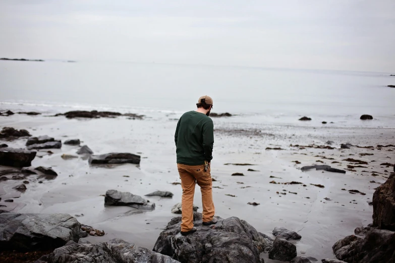 a man standing on top of a rock next to the ocean, a picture, unsplash, visual art, looking melancholy, stephen shore & john j. park, overcast day, hunched over
