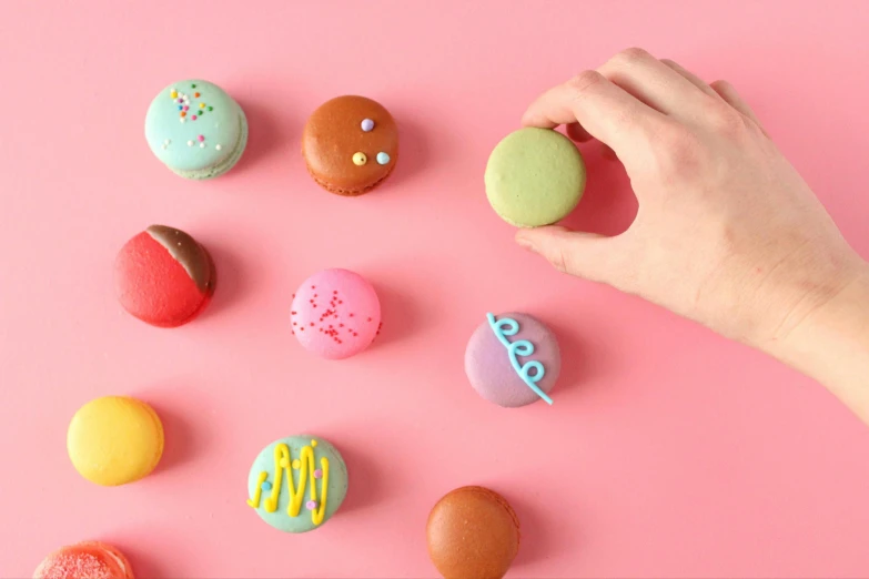 a person's hand reaching for colorful macarons on a pink surface, inspired by Damien Hirst, trending on pexels, 9 9 designs, clay animation, cupcake, postapo game