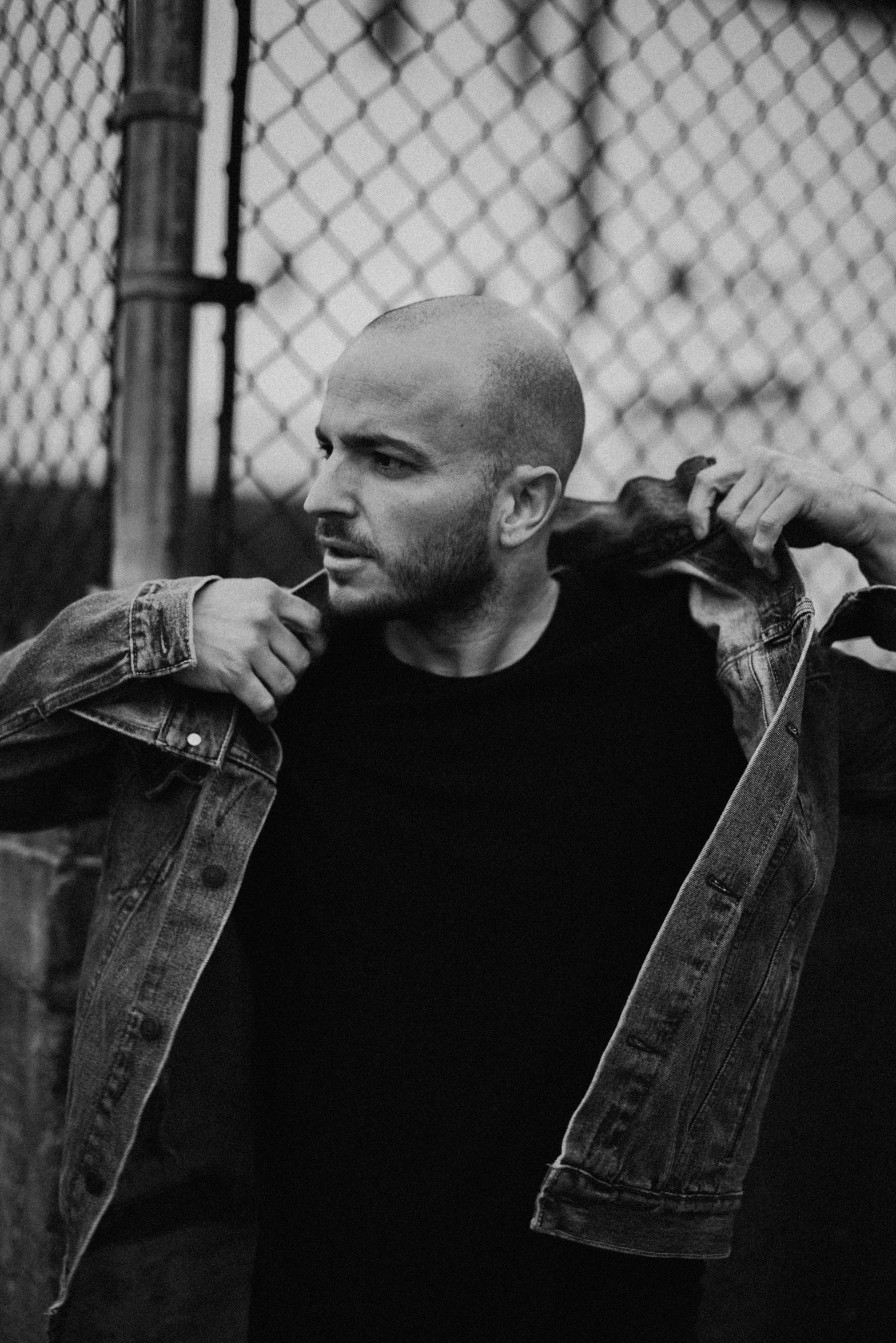 a man standing in front of a fence, a black and white photo, by Giorgio Cavallon, bald man, jean jacket, liam, ney matogrosso with malice