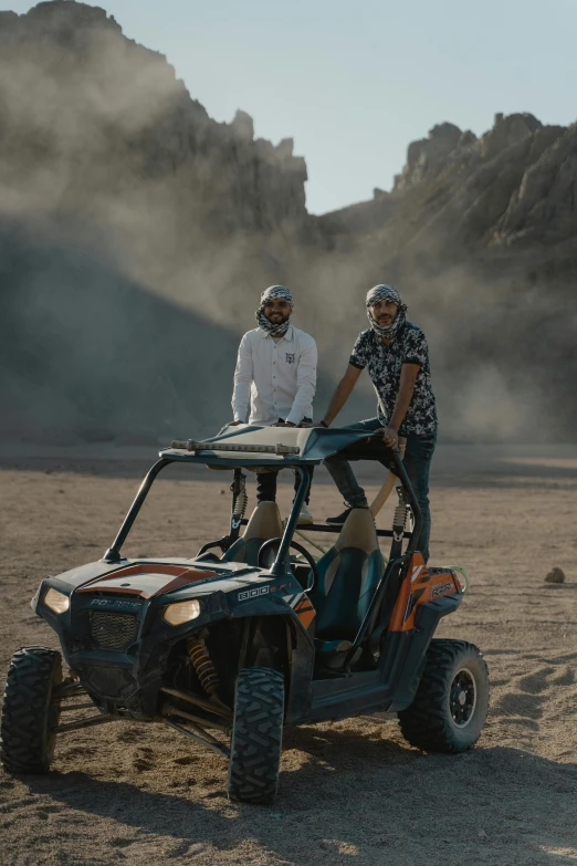 a couple of men riding on the back of a buggy, desert setting, bts, helmet is off, grey