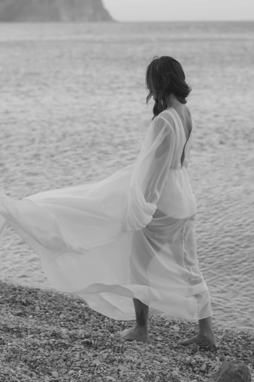 a woman standing on top of a beach next to a body of water, a black and white photo, romanticism, nico wearing a white dress, wearing a long flowy fabric, sheer fabrics, standing with her back to us