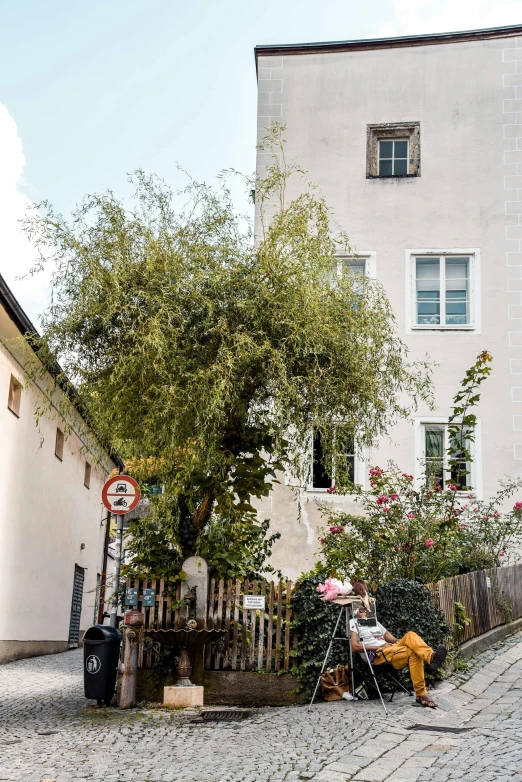 a person sitting on a bench in front of a building, arbeitsrat für kunst, giant white tree, narrow street, overgrown greenery, austrian architecture