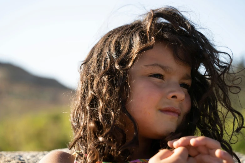 a little girl sitting on top of a rock, brown curly hair, in the sun, markings on her face, seeds