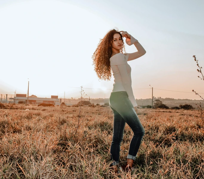 a woman standing in the middle of a field, by Marshall Arisman, pexels contest winner, happening, brown curly hair, jeans and t shirt, amazing beauty, wearing tight simple clothes