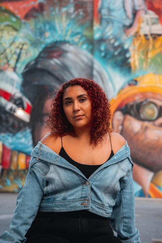 a woman standing in front of a graffiti wall, visual art, curly copper colored hair, hispanic, wearing double denim, center of image